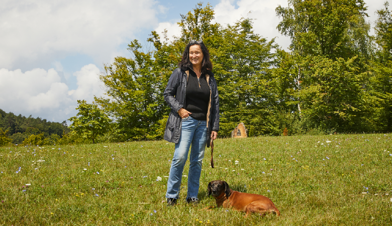 Eine Frau mit steht auf einer Wiese, vor ihr liegt ein Hund. 