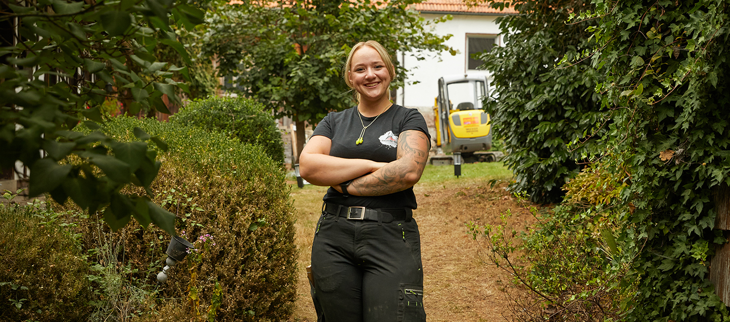 Eine Frau in Handwerksleidung steht mit verschränkten Armen in einem Garten vor einem Haus.