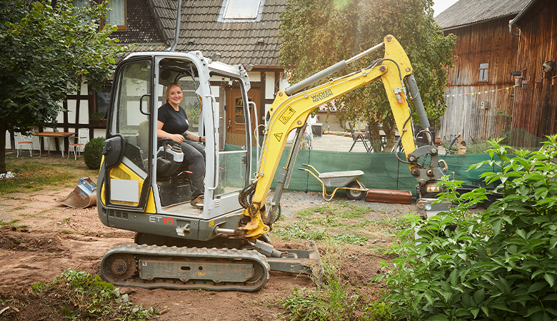 Eine Frau sitzt m Fahrzeughaus eines kleinen Baggers und hebt Erde im Garten aus.