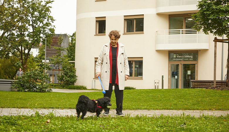 Eine Frau steht mit ihrem Hund in einer Grünanlage vor einem Haus.