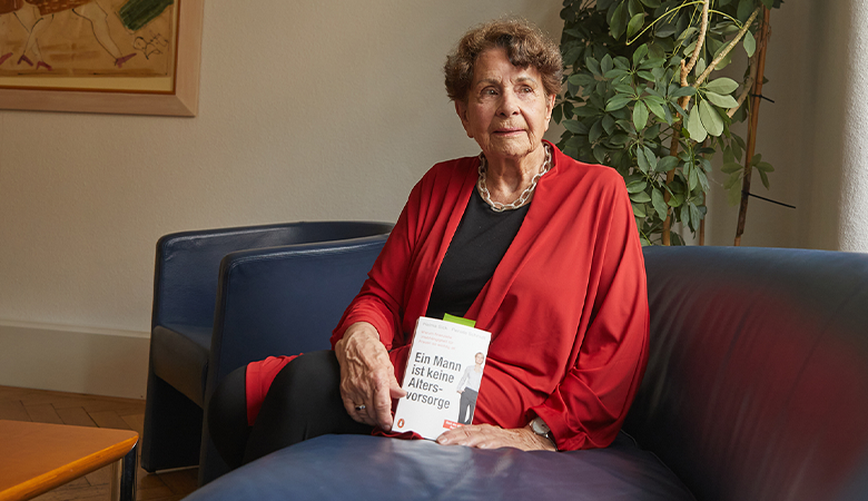 Eine Frau sitzt auf einem Sofa und hält ein Buch mit dem Titel "Ein Mann ist keine Altersvorsorge" in der Hand. 