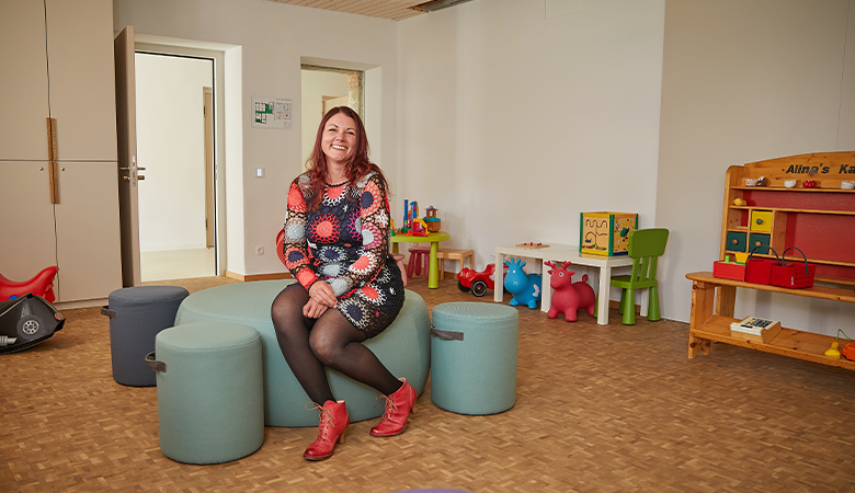 Eine Frau sitzt auf einem Sitzmöbel in einem Raum mit Kindermöbeln und Spielzeug. 