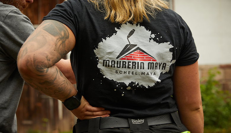 Rücken einer Frau mit einem T-Shirt, auf dem Maurerin Maya steht. 