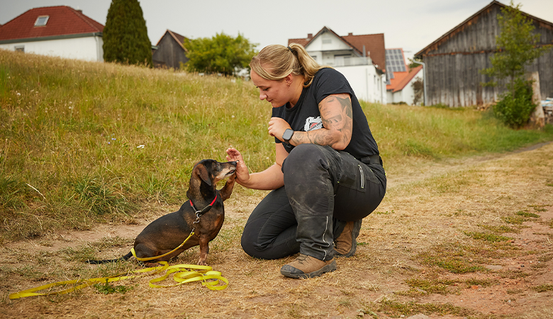 Eine Frau kniet auf einem Weg auf dem Boden. Vor ihr sitzt ein Dackel, der seine Pfote an ihre Hand hält. 