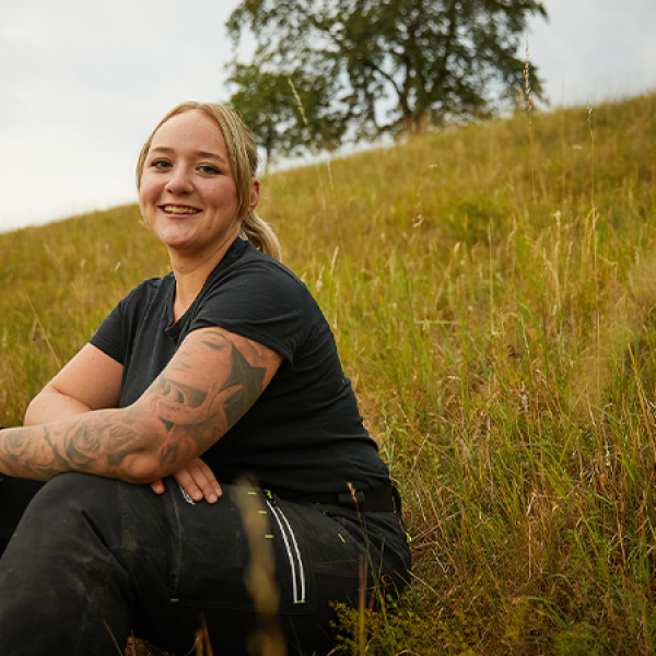 Eine Frau in Handwerkskleidung sitzt auf einer hügeligen Wiese und lächelt. 