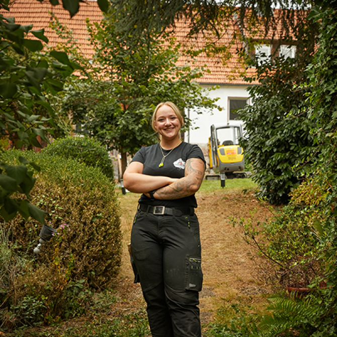Eine Frau in Handwerksleidung steht mit verschränkten Armen in einem Garten vor einem Haus.