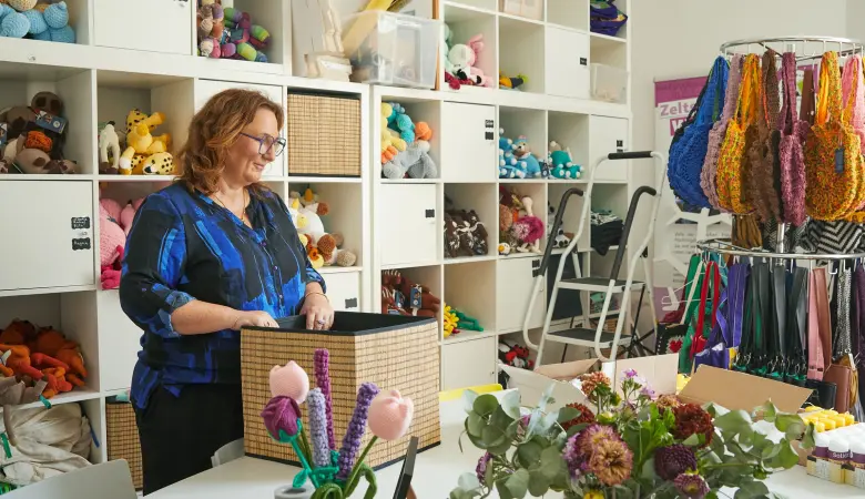 Eine Frau steht an einem Tisch und packt eine Kiste. Hinter ihr ist ein Regal mit Stricksachen. 
