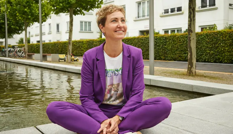 Eine Frau sitzt im Schneidersitz auf einem Holzsteg an einem Fluß in der Stadt.