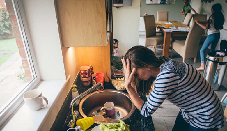 In der Küche: Eine Frau beugt sich genervt über den Abwasch. Im Hintergrund toben ihre Kinder.
