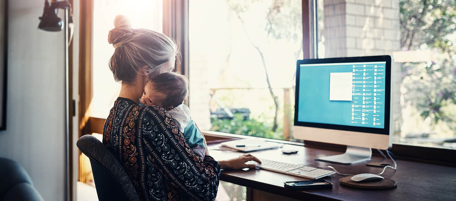 Im Homeoffice: Eine junge Frau arbeitet am Computer. Auf dem Arm hält sie ein Baby.
