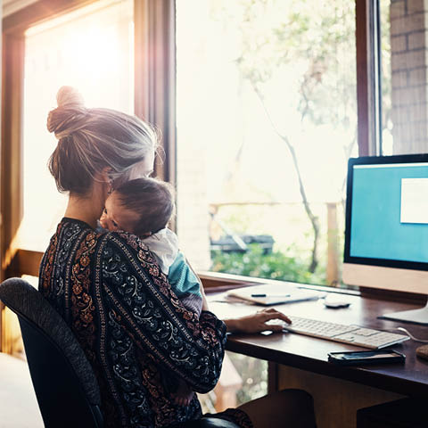 Im Homeoffice: Eine junge Frau arbeitet am Computer. Auf dem Arm hält sie ein Baby. 