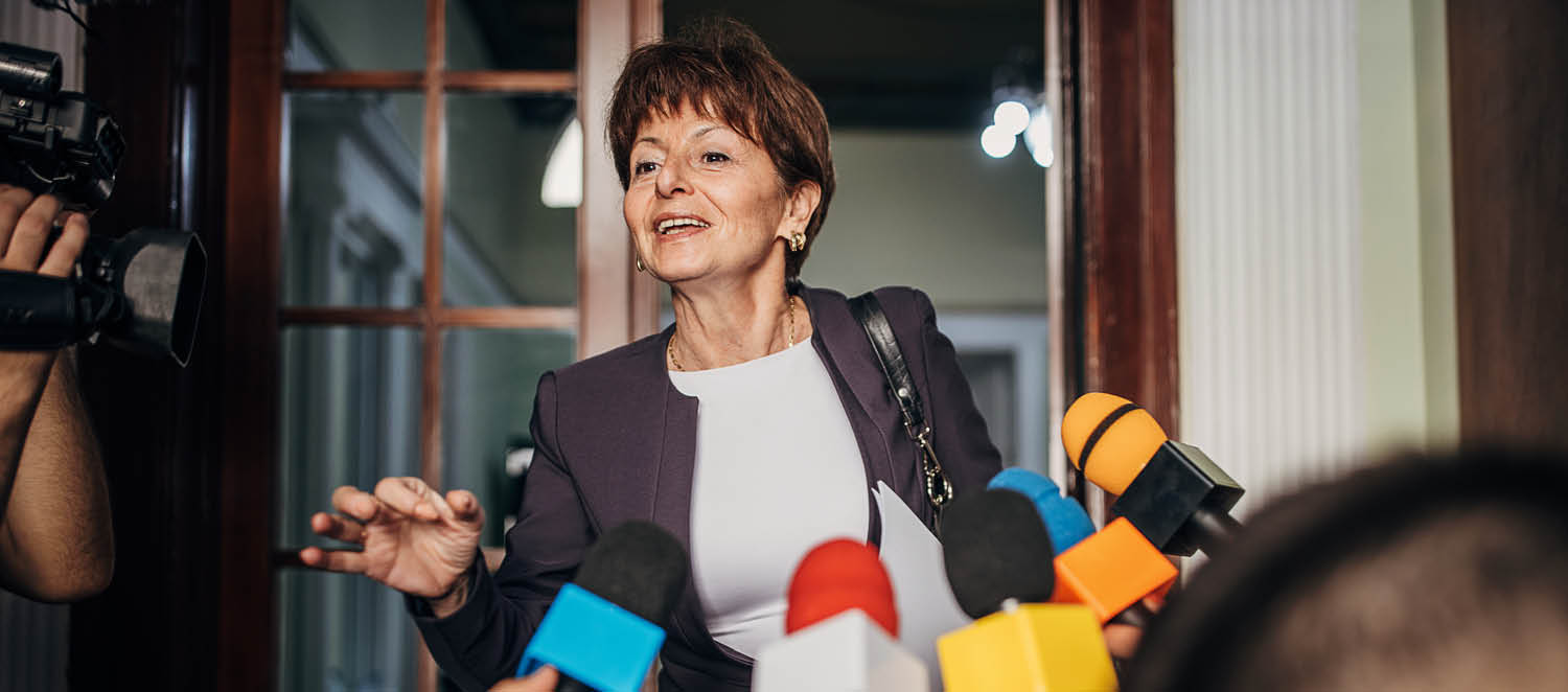 Eine Frau spricht und gestikuliert mit der Hand. Es sind Mikrofone und eine Kamera auf Sie gerichtet. 