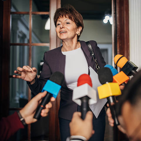 Eine Frau spricht und gestikuliert mit der Hand. Es sind Mikrofone und eine Kamera auf Sie gerichtet.