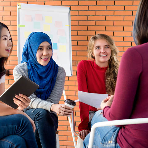 In einem Raum mit Flipchart: Eine diverse Gruppe junger Frauen sitzt im Kreis und diskutiert angeregt. 