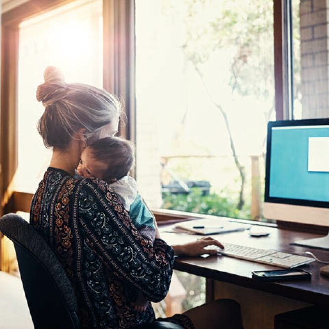 Im Homeoffice: Eine junge Frau arbeitet am Computer. Auf dem Arm hält sie ein Baby.