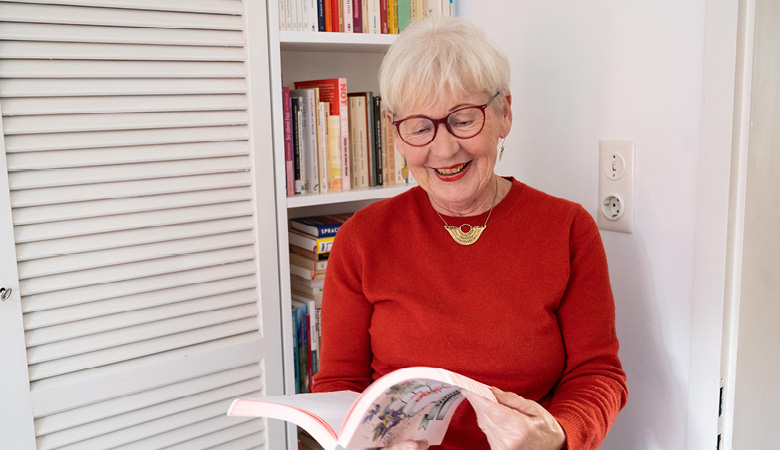 Elisabeth Brock steht vor einem Bücherregal. Sie hält ein großes, modern gestaltetes Buch in den Händen. Sie lächelt.