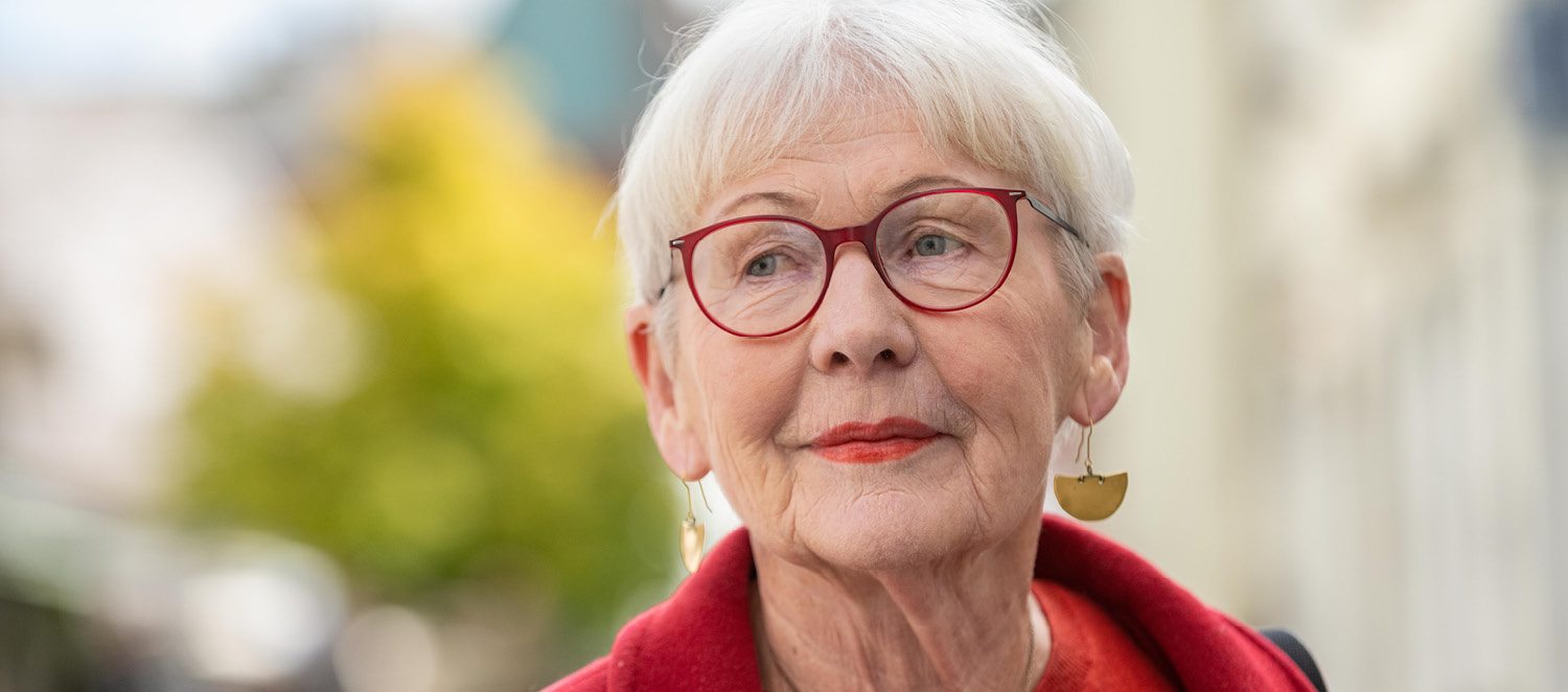 Porträtfoto: Elisabeth Brock. Sie hat weiße, kurz geschnittene Haare und trägt Brille, Pulli und Wollmantel in Rot.