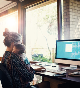 Im Homeoffice: Eine junge Frau arbeitet am Computer. Auf dem Arm hält sie ein Baby.