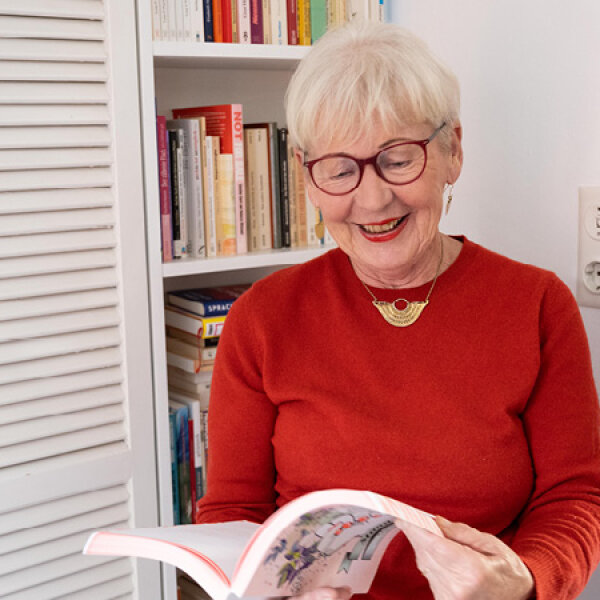 Elisabeth Brock steht vor einem Bücherregal. Sie hält ein großes, modern gestaltetes Buch in den Händen. Sie lächelt.