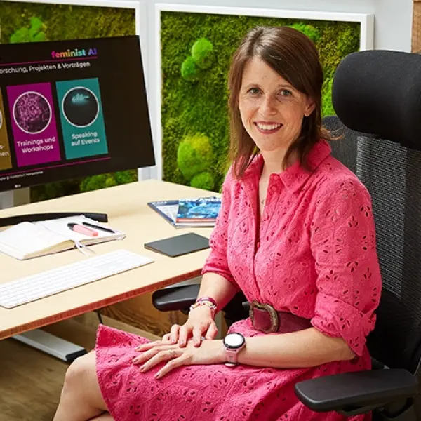 Eine junge Frau sitzt an einem Tisch mit einem Computer und lächelt in die Kamera.