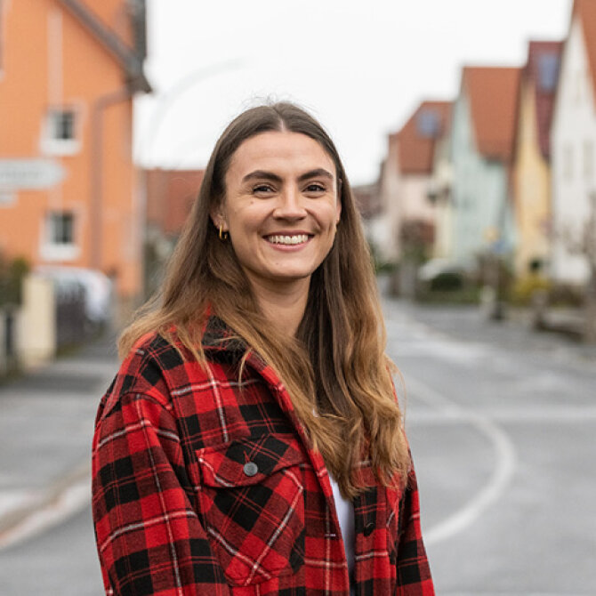 Porträtfoto: Lisa-Marie Schmitt steht an einer Dorfstraße, in der sich Einfamilienhäuser aneinanderreihen. Sie hat lange braune Haare und trägt eine rotkarierte Jacke.