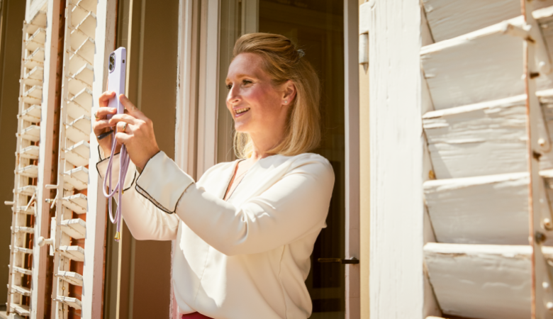 Claudia Alfons steht an einem geöffneten Fenster. Sie blickt in die Kamera Ihres Smartphones.