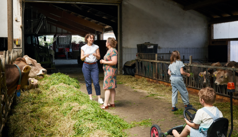 Zwei Frauen stehen in einem Kuhstall und unterhalten sich. Ein Junge fährt mit einem Kinderfahrzeug durch den Stall, ein Mädchen schaut zu den Kühen.  