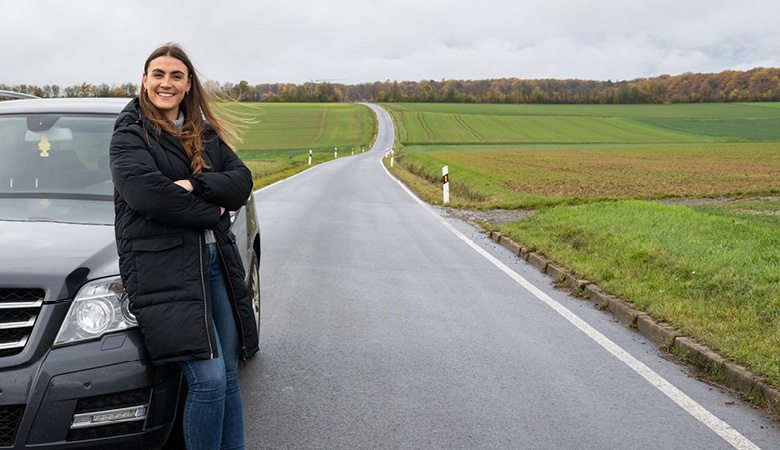 Auf einer schmalen Landstraße. Lisa-Marie Schmitt lehnt an ihrem Auto. Sie lacht. 