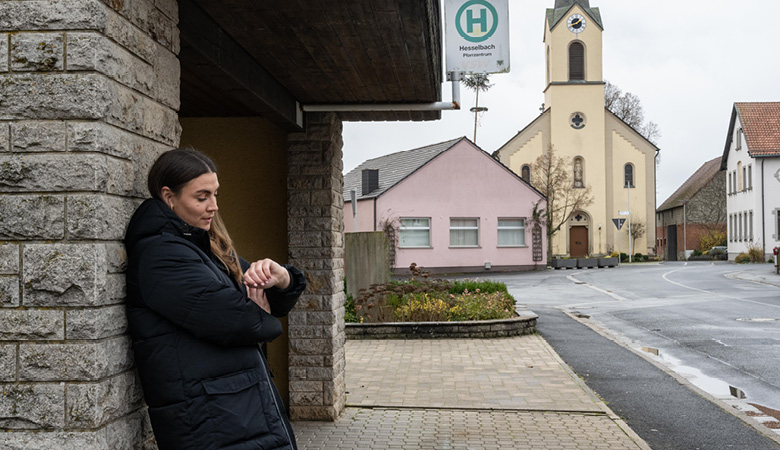 Lisa-Marie Schmitt steht vor einem gemauerten Buswartehäuschen. Sie schaut auf ihre Armbanduhr.