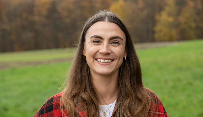 Porträtfoto: Lisa-Marie Schmitt steht in einer grünen Landschaft; im Hintergrund erstreckt sich ein Waldstück. 