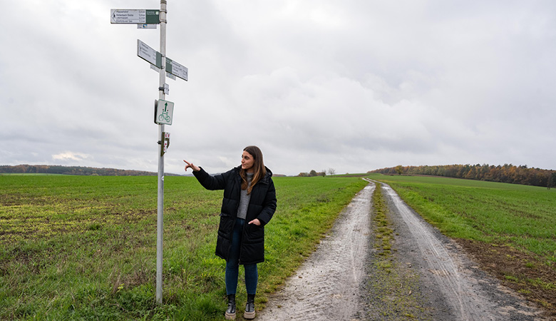 Lisa-Marie Schmitt steht am Rand eines Feldwegs neben einem Wegweiser mit mehreren Schildern.