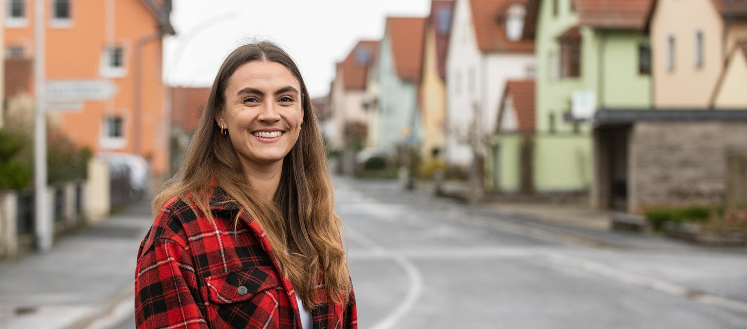 Porträtfoto: Lisa-Marie Schmitt steht an einer Dorfstraße, in der sich Einfamilienhäuser aneinanderreihen. Sie hat lange braune Haare und trägt eine rotkarierte Jacke.