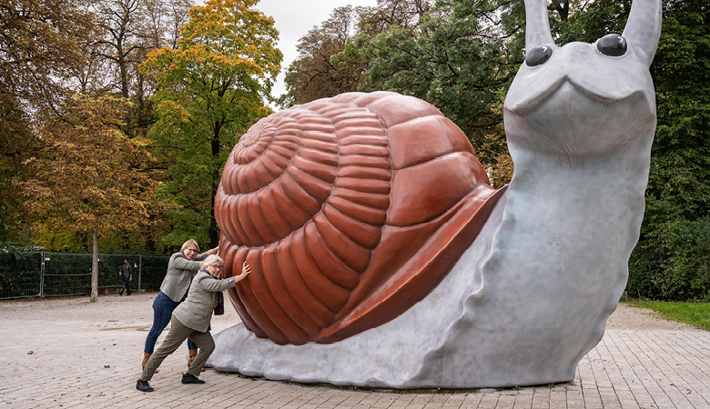 Marianne Schweinesbein und Karin Stempfhuber stehen vor einer mehr als vier Meter hohen Schnecken-Skulptur. Sie drücken gegen das Schneckenhaus, als wollten sie das Tier anschieben.