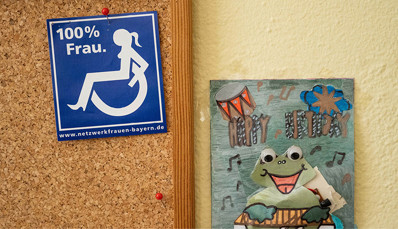 Nahaufnahme: eine Pinnwand mit einem Aufkleber. Er zeigt das Symbol einer Frau im Rollstuhl.