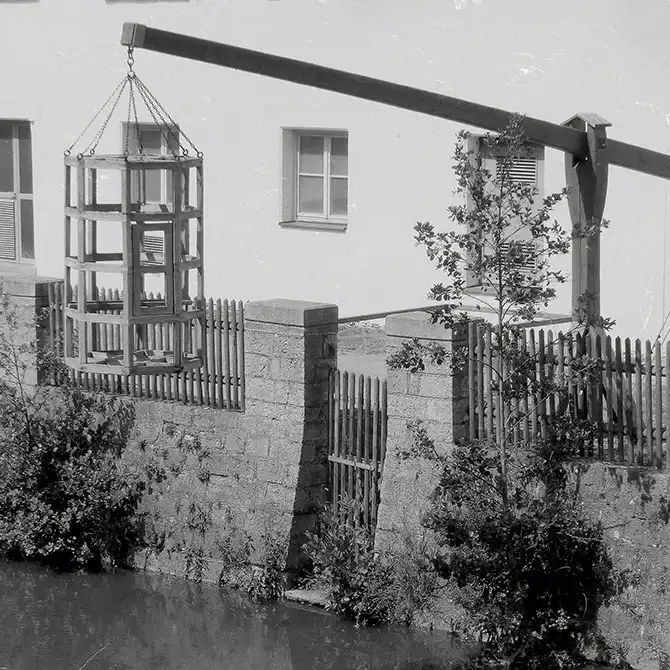 Ein Holzkäfig, der vor einem Haus über einem Fluss hängt.